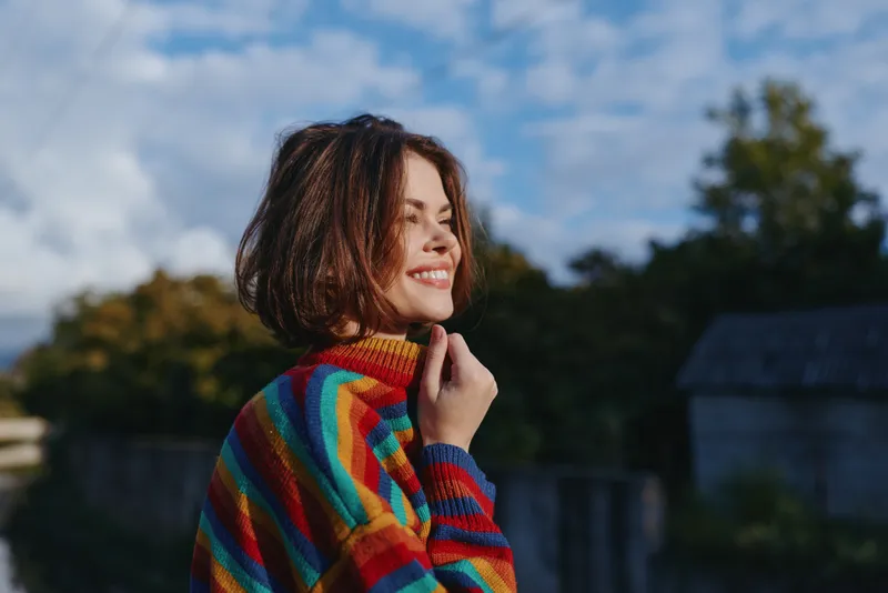 Woman smiling in a colorful striped sweater outdoors, young adult with short hair enjoying countryside breeze, casual outfit and relaxed lifestyle portrait for fashion, travel and mood.