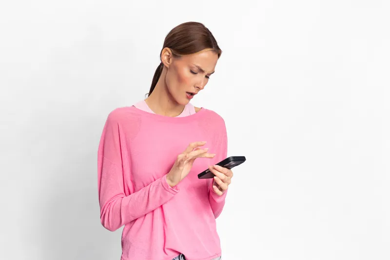 Young woman in pink sweater looks surprised at smartphone screen isolated on white. Concept for communication, technology, emotions, and digital lifestyle.