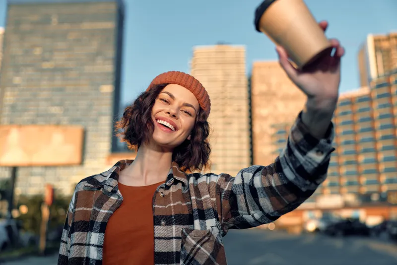 Woman holding coffee cup in urban street, smile and candid lifestyle moment with beanie and plaid shirt, sunlight and happiness combine into golden hour glow, authenticity and mindful living.