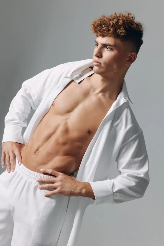 Young man with curly hair posing shirtless in white pants and open shirt, showing toned abs and muscular body against plain gray background in studio.
