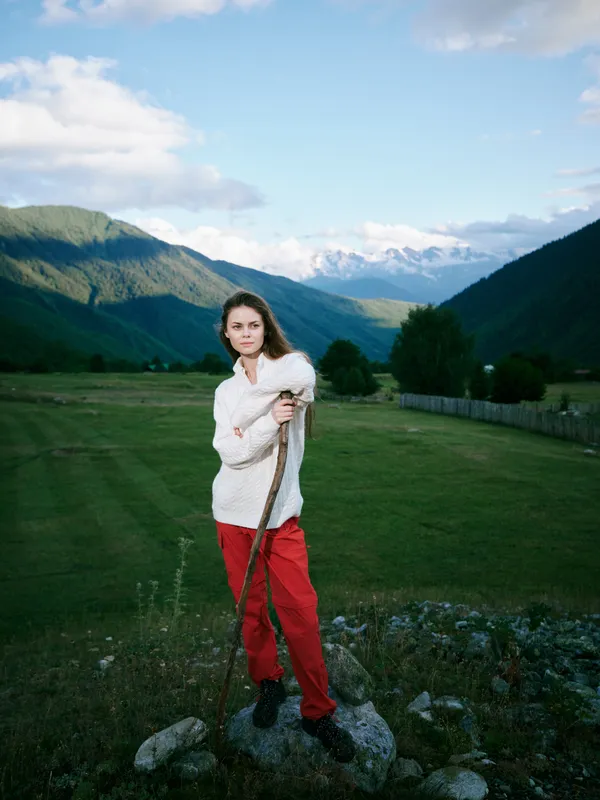 Landscape with distant mountains, a woman in a light coat standing on a grassy field, holding a wooden staff in a serene outdoor scene, emphasizing nature and calm weather