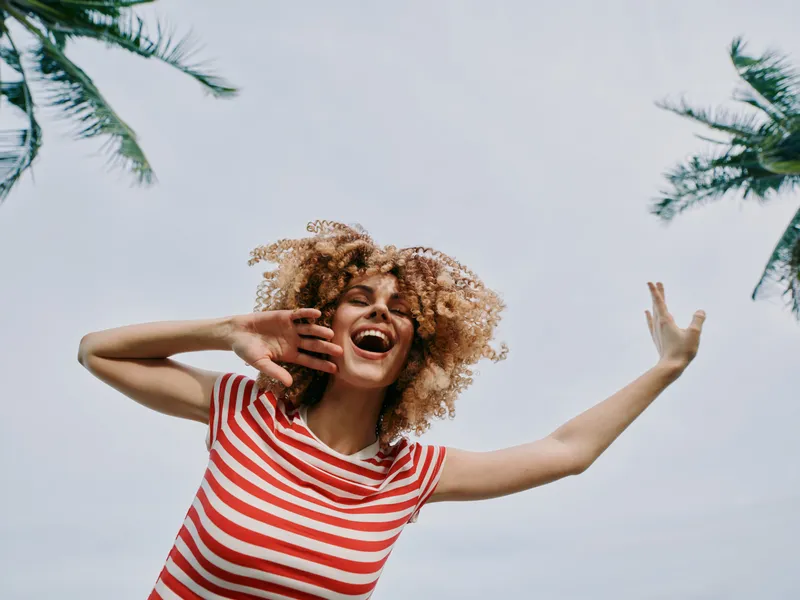 Vibrant candid portrait of a joyful young woman with curly hair in a red striped shirt, laughing and dancing under palm trees against a clear bright sky, exuding carefree energy and sunshine.