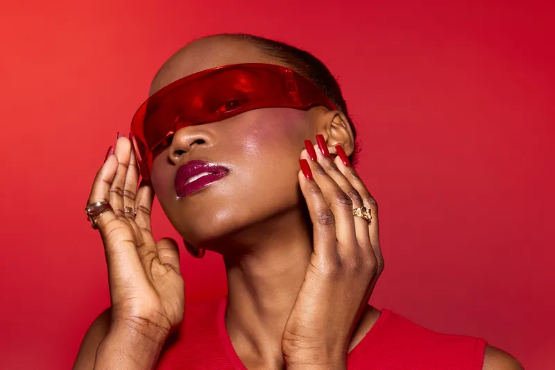 Stylish dark skinned African woman with shaved head wearing red visor and red nail polish poses on red background. Concept of fashion, beauty, confidence, and modern style.