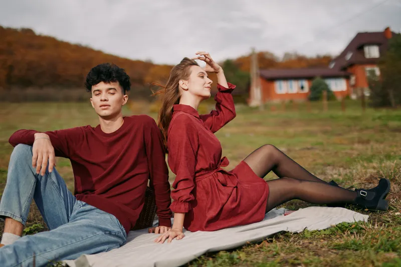 Young couple sits back to back on a picnic blanket in a rural field, natural poses and subtle smiles conveying authenticity and credibility in a quiet, candid lifestyle moment.