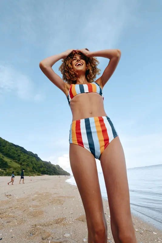 A happy young woman in a colorful striped bikini stands on a sandy beach near the water with green hills in the background under a clear blue sky.