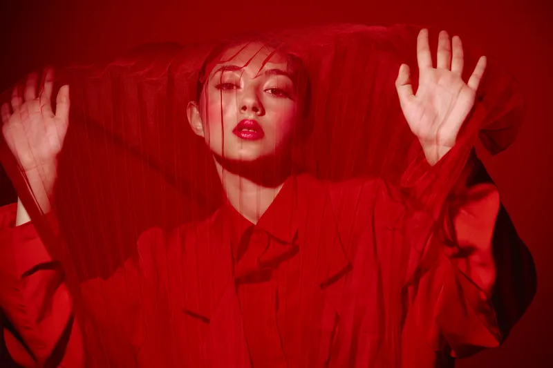 Fashionable young woman in red outfit behind red mesh fabric. Creative beauty concept with strong color contrast on a red background. Artistic high fashion studio shot.