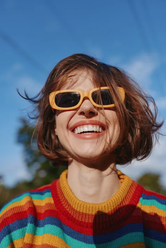 Smiling woman wearing sunglasses and colorful sweater, short hair portrait outdoors in sunlight. Joyful young adult enjoying bright day, candid closeup with natural background and vibrant tones.