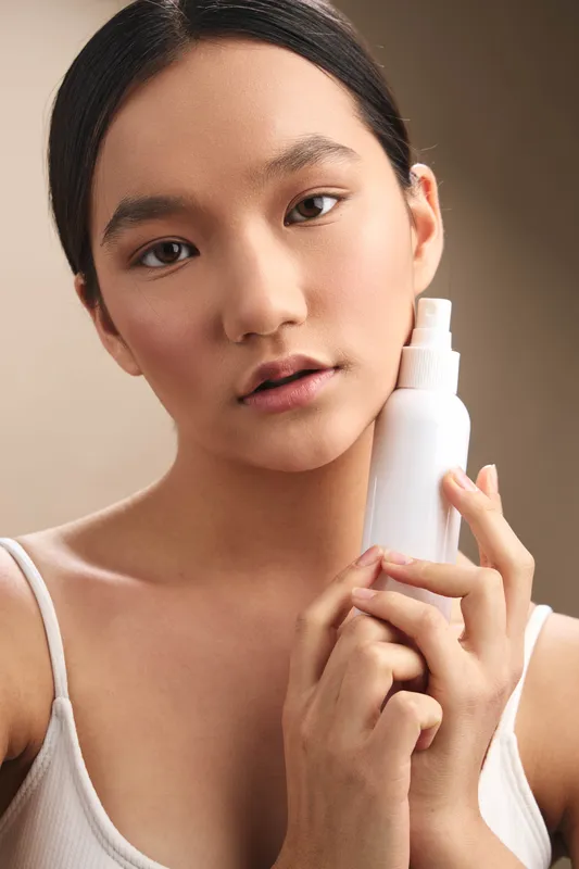 Asian woman is pictured in a clean studio environment, holding a skincare bottle near her face with a soft expression, conveying calm confidence and self care.