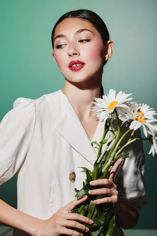 Young woman in white blazer holding white daisies looks sideways with glossy red lips and styled hair. Soft lighting highlights her serene expression in fashion beauty concept.