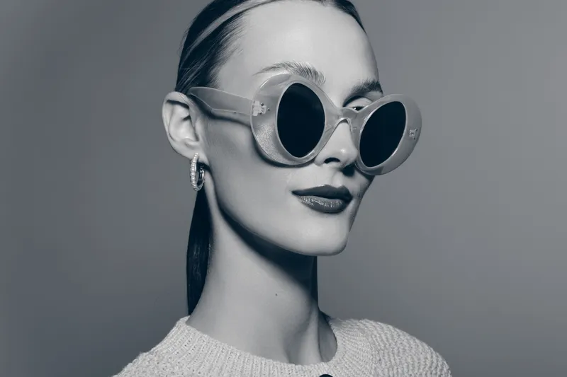 Young Caucasian woman with fair skin wearing round sunglasses and earrings poses confidently in studio portrait.
