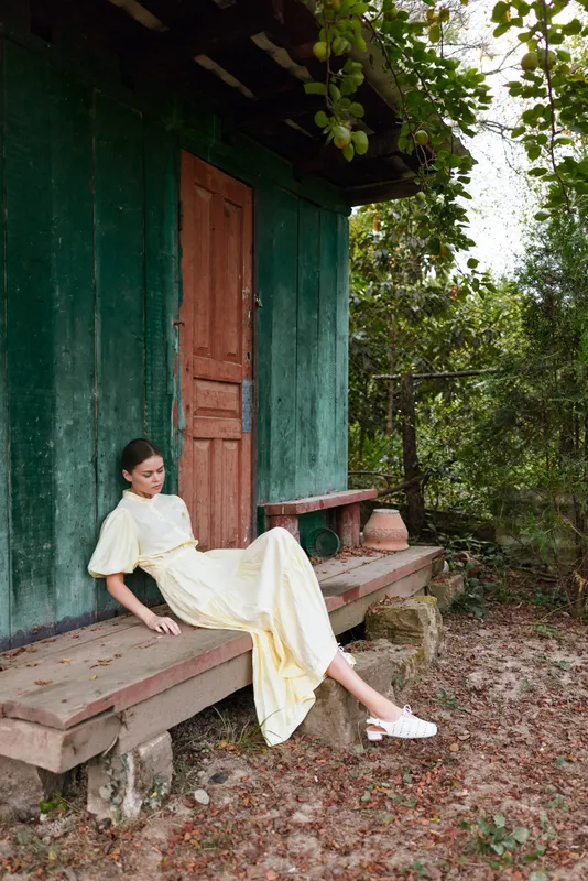 Woman in a pale yellow dress sits on the porch steps beside a green wooden house, relaxed pose in an outdoor rustic setting, sunlight filters through trees creating a calm timeless atmosphere