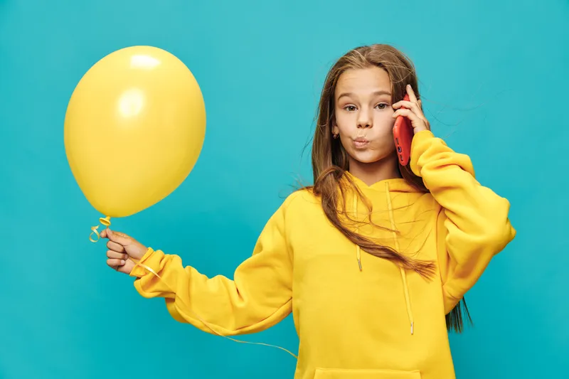 Girl in yellow hoodie holding yellow balloon and talking on red smartphone with blue background. Child with long hair expressing surprise or curiosity while communicating on phone.