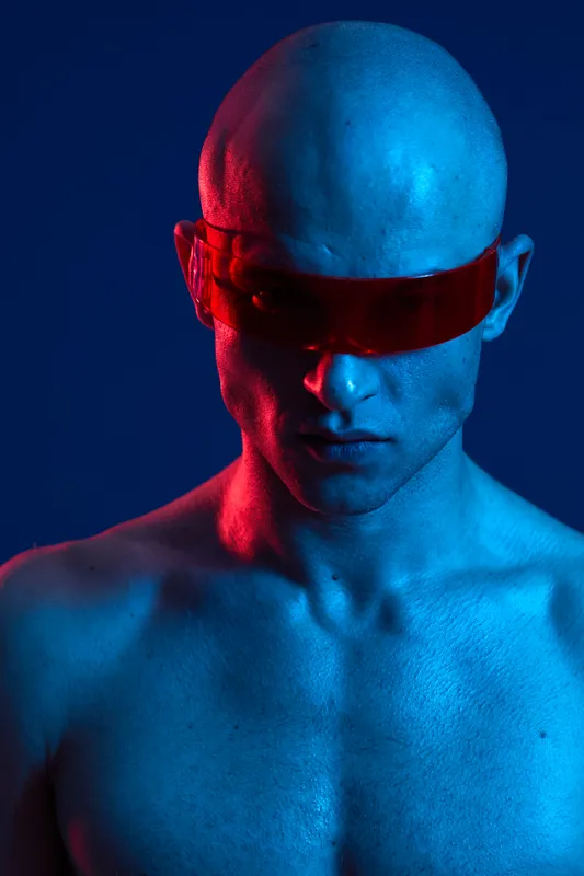 Striking man with futuristic sunglasses in blue and red lighting, presenting a dynamic and bold character. The image captures a blend of modern style and attitude.