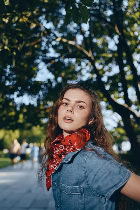 young woman with red scarf and denim shirt standing outdoors under lush green trees during daytime, natural light and casual summer style