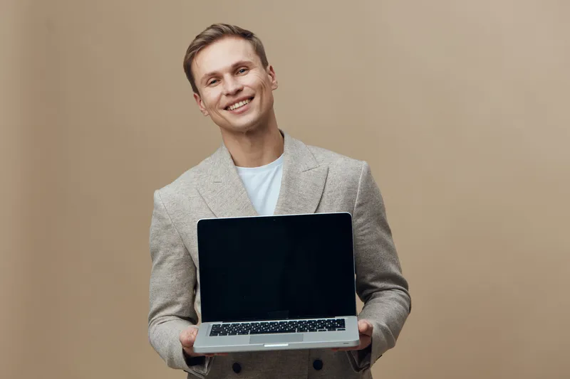 Smiling young Caucasian man in light blazer holding open laptop computer, looking cheerful and confident, standing against plain beige background, lifestyle concept.