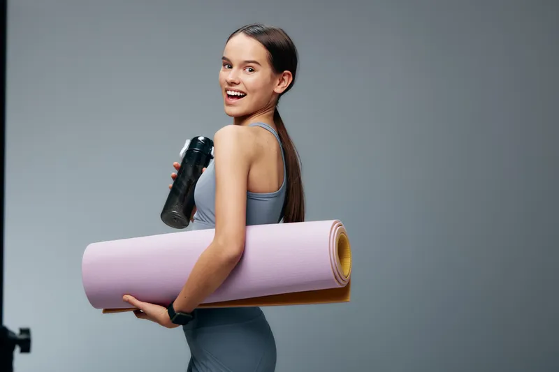 Active woman in athletic wear carries a pink yoga mat and water bottle, ready for a workout. Gym environment, focus on fitness, motivation and training routine.