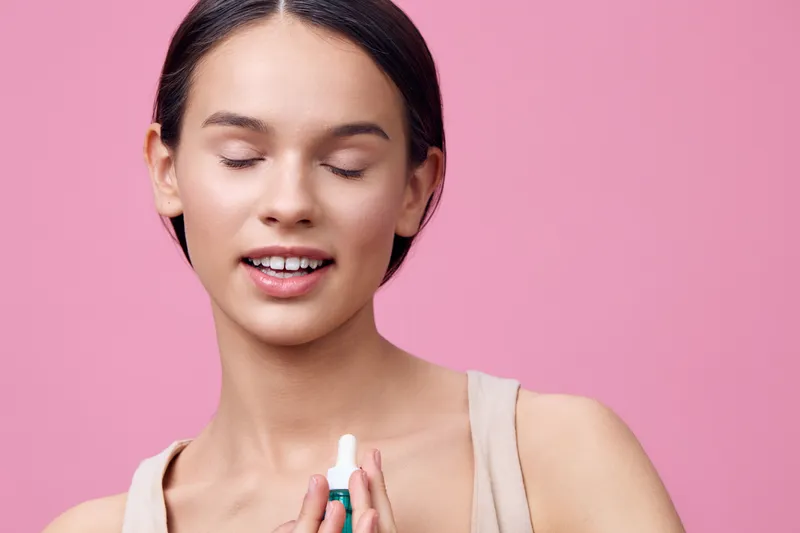 Young woman showcasing a skincare product with closed eyes, smiling against a pink background, promoting self care and healthy skin routine