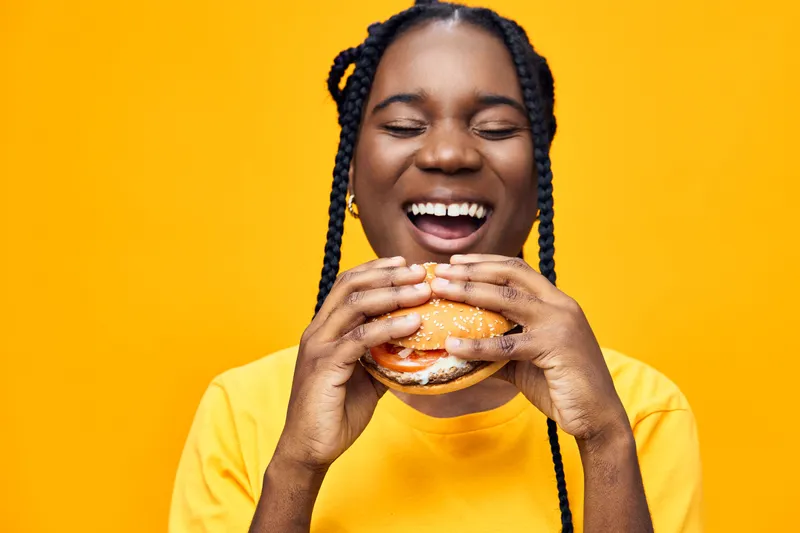 Cheerful African woman enjoying a delicious burger against a vibrant yellow background, wearing a bright yellow t shirt, embodies happiness and healthy eating