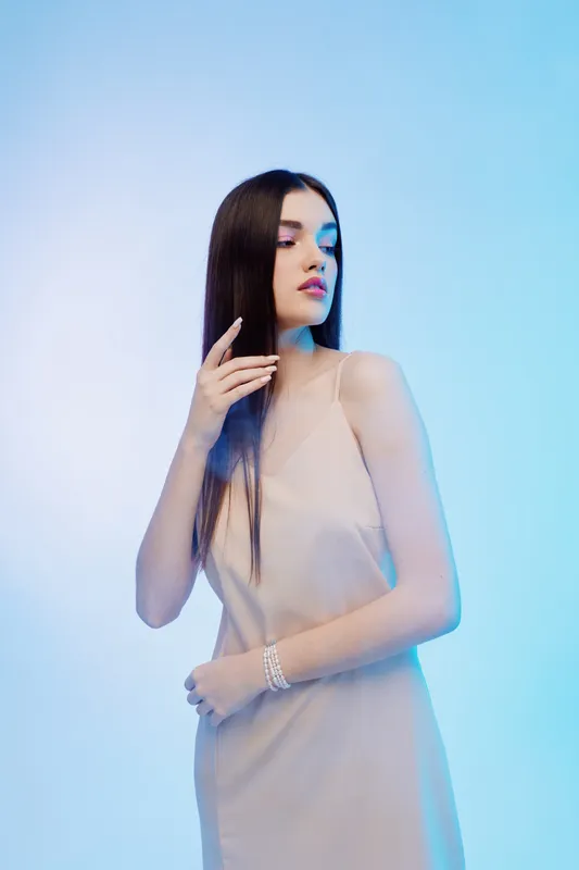 Young woman with long dark hair poses in a beige dress against a blue background. Concept of beauty, fashion, elegance, and modern style in studio lighting.