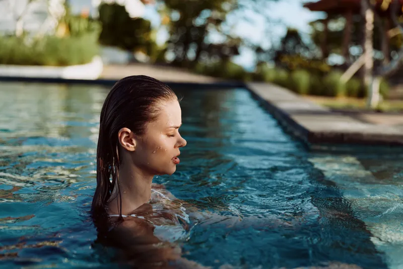 Young woman with wet hair relaxes in outdoor swimming pool during sunny day surrounded by trees and clear water lifestyle concept