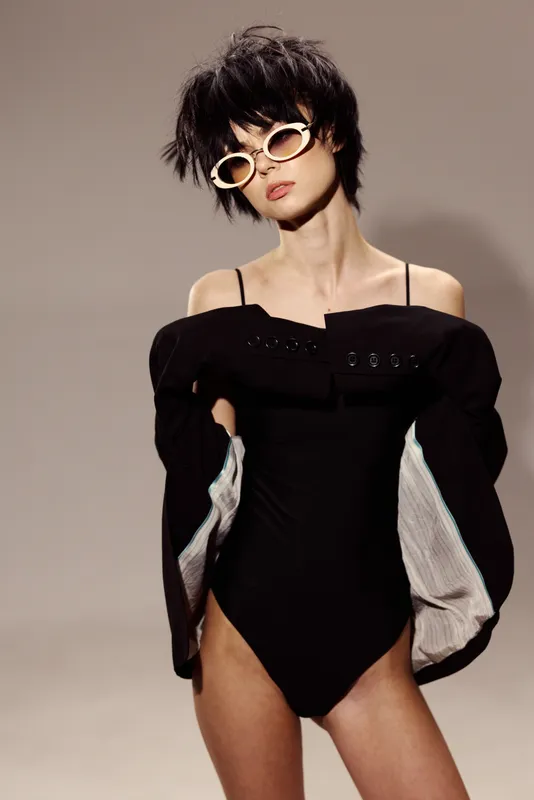 Trendy woman in black bodysuit and sunglasses posing confidently with tousled hair, stylish and modern fashion.