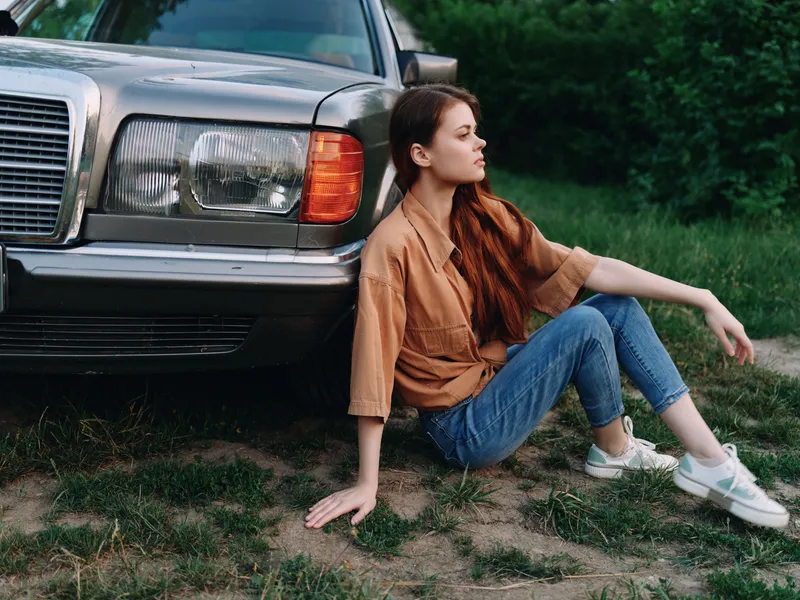 Young woman sitting near an old car, showcasing relaxed vibes in a natural green setting, dressed casually in a beige shirt and jeans, depicting freedom and nostalgia.
