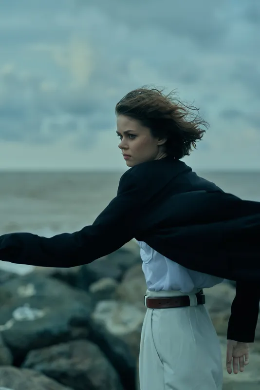Windy seaside scene with a woman in a white dress and dark cape, standing on rocks by the ocean, facing the wind with a determined gaze, conveying resilience, movement and coastal fashion.