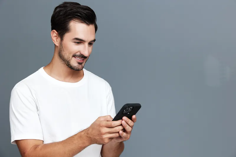 Smiling man with short dark hair wearing a white t-shirt, happily using a smartphone while standing against a soft gray background in a studio portrait.