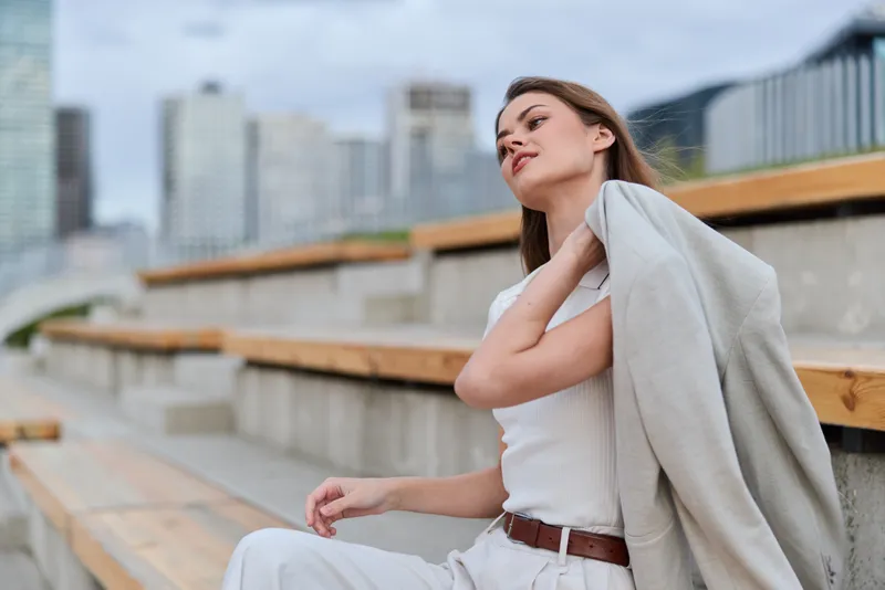 Young woman in casual white outfit holding jacket outdoors with urban cityscape background, thoughtful emotion, spring style fashion photo