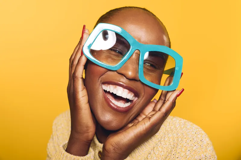 Happy African woman with dark skin wearing oversized blue glasses and yellow sweater smiles brightly on yellow background. Concept of joy, fashion, and vibrant lifestyle portrait.