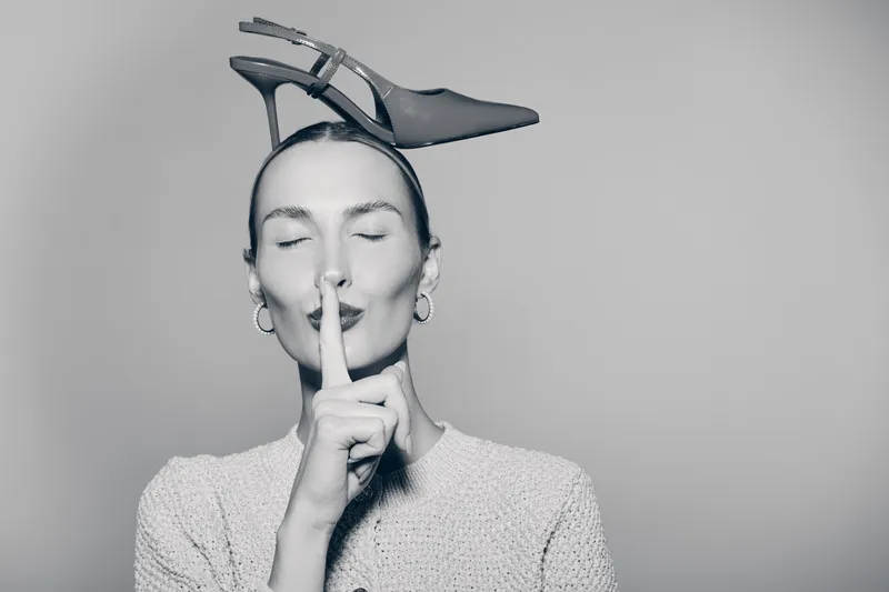 Young white woman with pale skin holding finger to lips and wearing high heel shoe on head in studio.