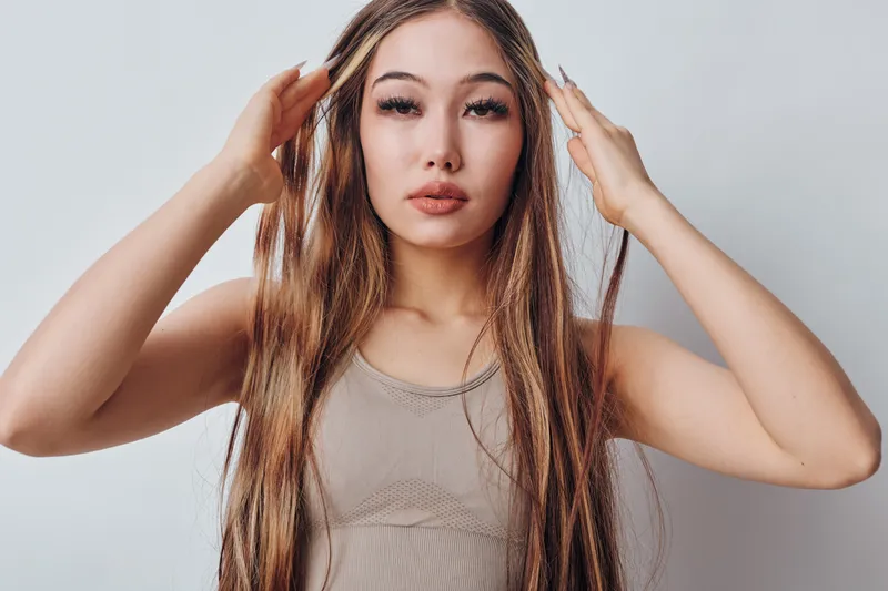 Young woman with long hair posing indoors with hands on head, looking confidently at camera. Beauty, fashion and portrait with natural makeup on neutral background.