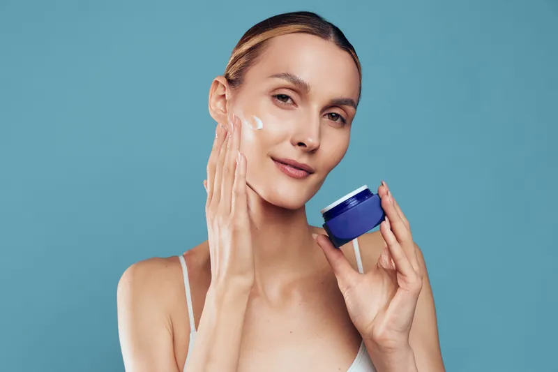 Woman applying face cream in spa skincare routine, holding blue cream jar, soft smile, self care beauty, smooth skin, studio light, blue background, healthy glow