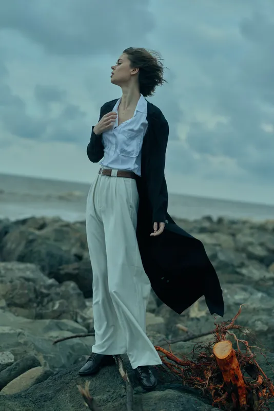 Elegant woman in a blazer and wide trousers stands on a rocky shore, wind in her hair, facing the horizon in a dramatic coastal fashion moment.