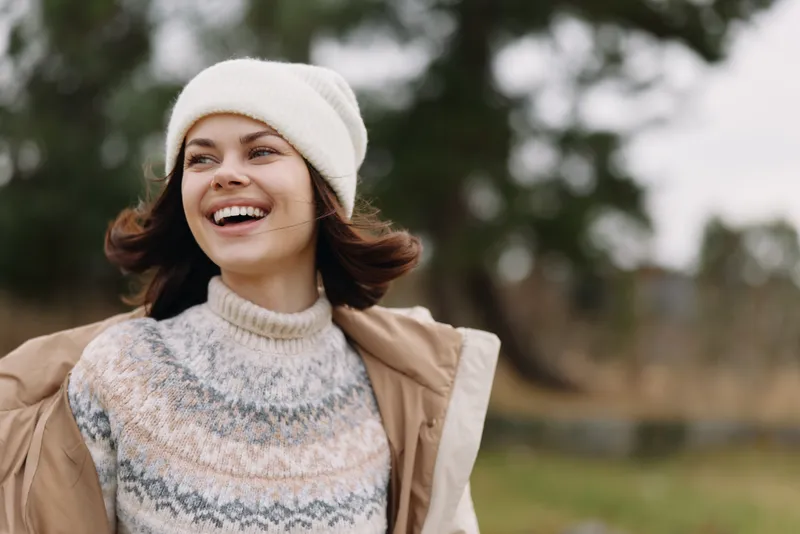 Woman, smiling, outdoor portrait featuring a cozy knit sweater and beanie, light jacket, carefree mood in a park setting during chilly weather