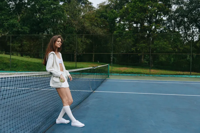 Young female tennis player in a white sporty outfit posing confidently on the tennis court with vibrant green background, showcasing athleticism and determination