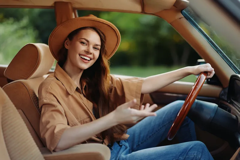 Cheerful young woman driving a vintage car, wearing a stylish hat and casual outfit, enjoying a sunny day in nature with a smile.
