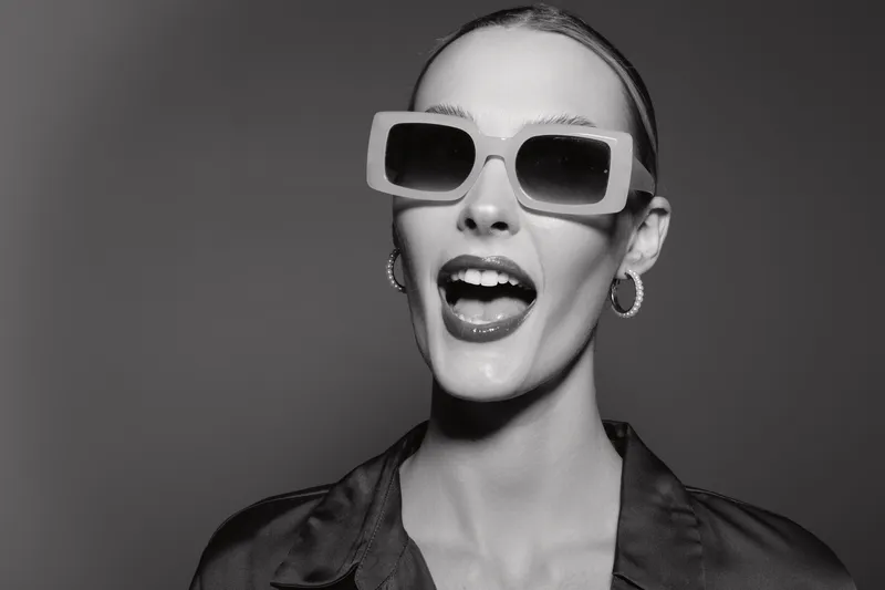 Portrait of a young white woman wearing oversized sunglasses and hoop earrings, smiling with open mouth on grey background.