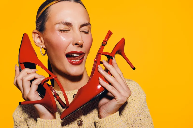 Happy woman with red lipstick holds red high heel shoes near face, eyes closed, smiling on bright yellow background. Concept of fashion, style, and joyful expression.