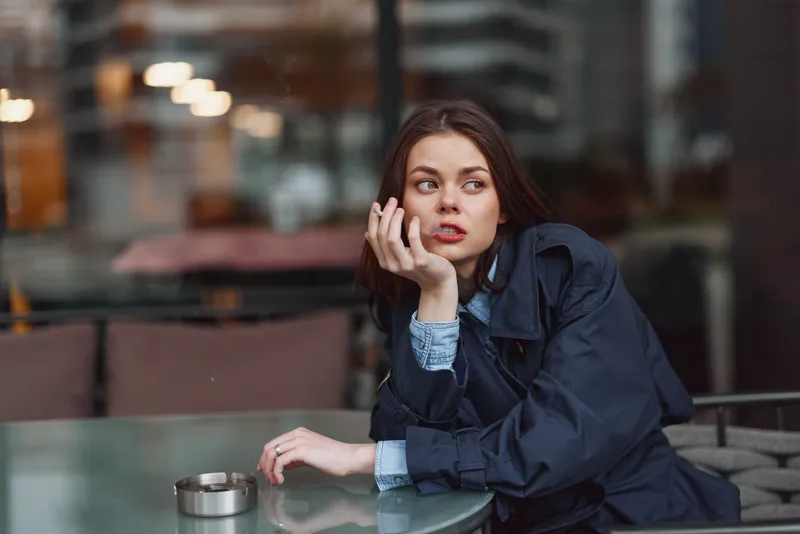 Young woman with long brunette hair wearing a dark coat sits thoughtfully at an outdoor cafe table with a glass ashtray nearby during an overcast day.