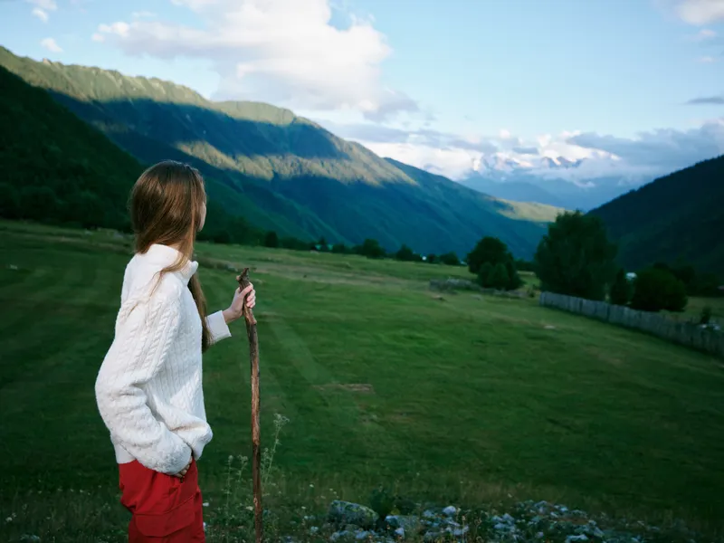 Woman in a white sweater with red pants stands on a hillside with a walking staff, overlooking a valley and distant mountains, capturing hiking and landscape serenity
