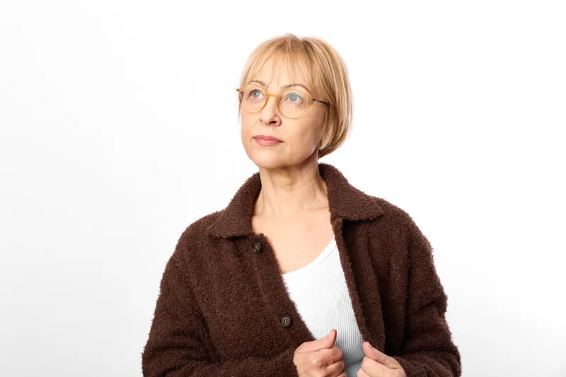 Senior woman with glasses in studio portrait, blond short hair, wearing brown coat and white top, calm expression, looking at camera, relaxed posture, soft even lighting