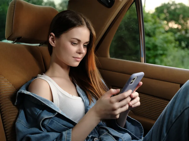 Young woman in casual denim attire using a smartphone in a car, showing a relaxed expression while gazing at the device, with a green nature background.