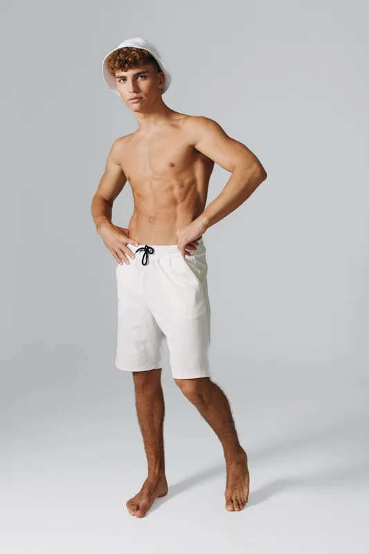 Young man with muscular body wearing white shorts and bucket hat standing barefoot on gray background. Fitness, summer style and casual fashion with confident pose and toned skin.