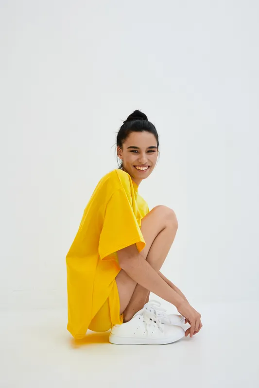 Smiling woman with dark hair in a yellow oversized t-shirt and white sneakers sitting on the floor, radiating youthful energy against a plain white background.