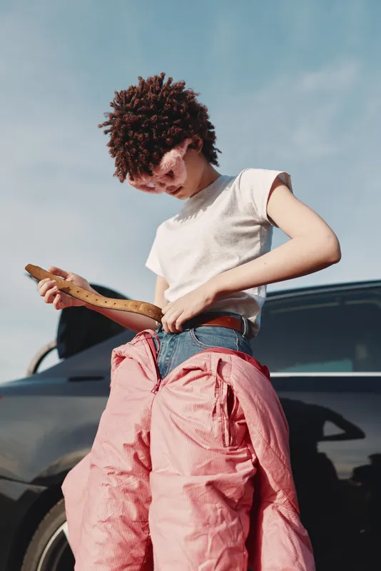 Street style, outdoor fashion with a person in pink pants and white top examining a baguette beside a car on a sunny daytime street, creating an urban casual vibe.