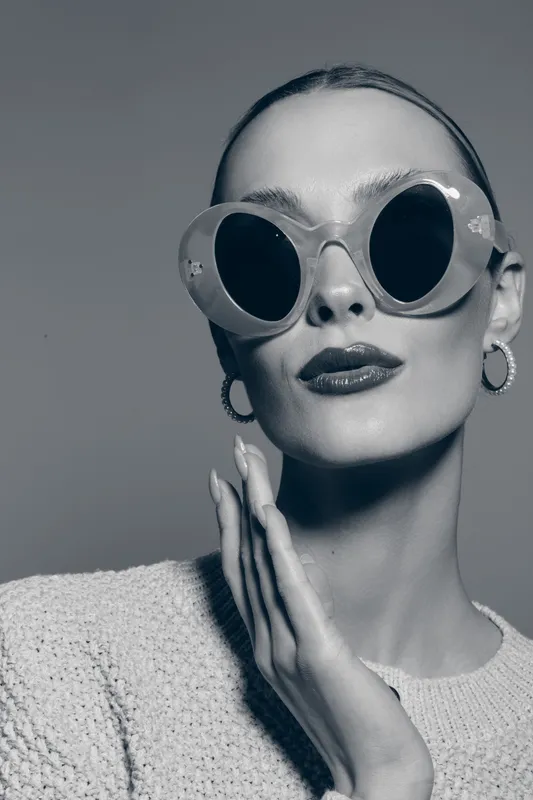Stylish white woman with fair skin wearing oversized round sunglasses and hoop earrings poses with hand near face in studio portrait.