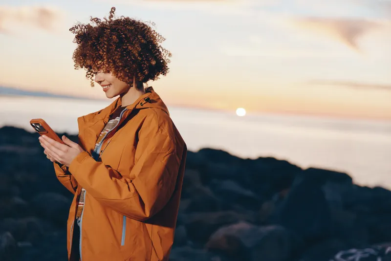 Woman with curly hair in a brown coat stands by a rocky beach, holding a phone and smiling softly. Golden sunset light and gentle wind suggest an outdoor lifestyle travel moment.
