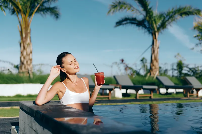 A relaxed woman in a white bikini enjoys a red cocktail while leaning on the edge of a pool with palm trees and lounge chairs in the background on a sunny day.