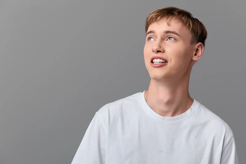Young man in a white tee gazes upward with a hopeful smile against a gray backdrop, conveying casual mood and everyday lifestyle appeal with bright, modern vibe